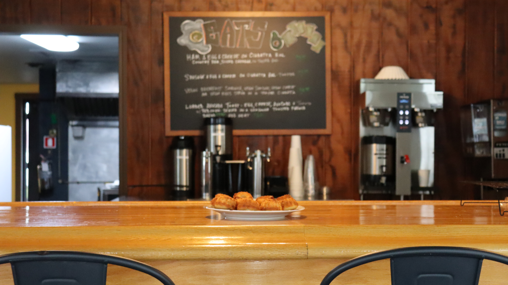 A plate of pastries sits on a wooden counter in a café, with a chalkboard menu and coffee machines blurred in the background.