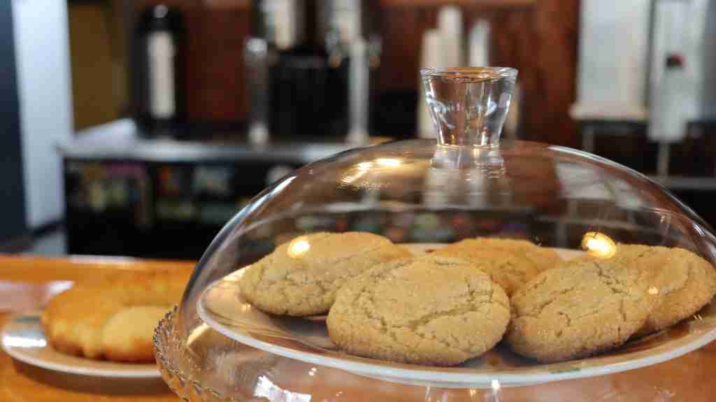 A plate of sugar cookies is displayed under a glass dome on a wooden counter, with a blurred background of a café and drink dispensers.