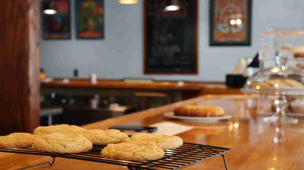 A batch of cookies cools on a wire rack on a wooden counter in a cozy café, with blurred pastries and colorful framed art in the background.