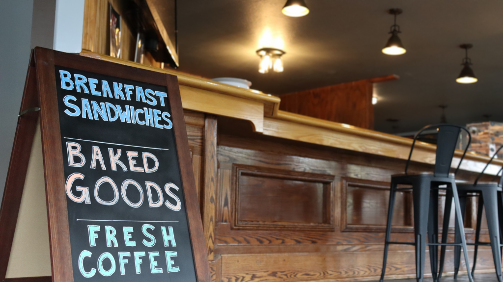 A chalkboard sign in front of a wooden counter displays Breakfast Sandwiches, Baked Goods, Fresh Coffee inside a cozy café with high chairs and warm lighting.